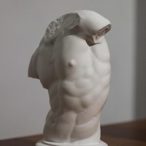 Pode incluir: Uma escultura de gesso branco de um torso masculino, sem cabeça nem braços. A escultura apresenta musculatura detalhada e é montada sobre uma base circular. A estátua é uma obra de arte clássica.