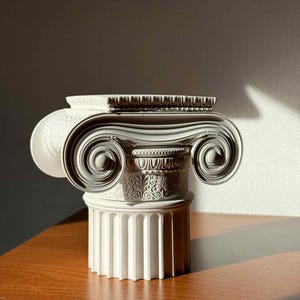 Op de afbeelding: Een witte, decoratieve Ionische kolomkapiteel met ingewikkelde krul- en bloemdetails. De kapiteel staat op een gefluteerde kolomvoet. Het architectonische element wordt weergegeven op een houten oppervlak, met een neutrale achtergrond.
