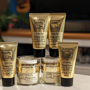 May include: A collection of gold cosmetic tubes and jars, labelled "Kindly, Joe" and "A-Hex-Pep Plumping Serum". The products are arranged on a white surface. The background is blurred. The tubes and jars are filled with a cream.