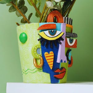 May include: An abstract face planter with a light green textured finish. The face design includes a blue eye, a yellow eye, a red mouth, and a yellow and red heart. The planter is filled with faux green and white plants.