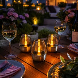 Puede incluir: Mesa al aire libre con tres faroles solares con forma de vela, copas de vino y una botella de vino. Los faroles tienen una carcasa de vidrio y una base metálica, creando un ambiente cálido y acogedor. La mesa es de madera.