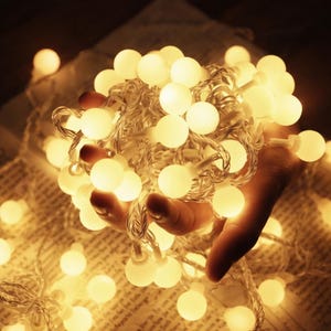 Puede incluir: Una mano sostiene una cadena de luces con bombillas blancas cálidas. Las luces están agrupadas, creando un brillo suave y acogedor. El cable es plateado.