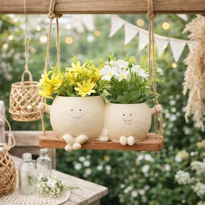 Puede incluir: Dos macetas de cerámica color crema con caras sonrientes, llenas de flores amarillas y blancas, se asientan en un columpio de madera. El columpio cuelga de una viga de madera con cuerda. Un encantador artículo de decoración de jardín.