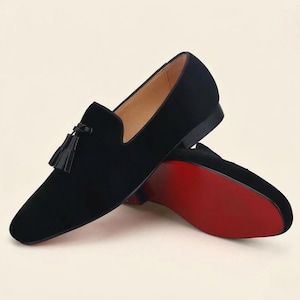 Könnte beinhalten: Ein Paar schwarze Wildleder-Slipper mit einer dekorativen Quaste auf dem Vorderblatt. Das Innenfutter ist hellbeige und die Außensohle ist leuchtend rot. Die Schuhe haben einen niedrigen Absatz und eine spitz zulaufende Form mit glatter Oberfläche.