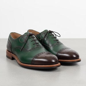 Könnte beinhalten: Ein Paar zweifarbige Lederschuhe. Die Schuhe sind grün mit braunen Zehenkappen und braunen Schnürsenkeln. Die Sohlen sind hellbraun. Die Schuhe sind mit dekorativen Perforationen versehen.