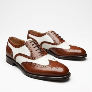 Könnte beinhalten: Ein Paar zweifarbige braun-weiße Leder-Wingtip-Schuhe. Die Schuhe haben ein klassisches Design mit aufwendigen Perforationen und dunkelbraunen Schnürsenkeln. Vor weißem Hintergrund.