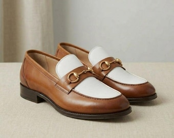 May include: A pair of two-tone loafers featuring a tan leather body and a white leather vamp. Each shoe is accented with a gold-toned metal horsebit detail across the top, a low stacked heel, and a rounded toe silhouette.