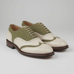 Könnte beinhalten: Ein Paar zweifarbige Oxford-Schuhe mit olivgrünen Wildledereinsätzen und cremefarbenen Lederpartien. Die Schuhe verfügen über dekorative Brogue-Perforationen entlang der Flügelkappe und der Nähte, beigefarbene Schnürsenkel sowie einen dunkelbraunen Absatz.