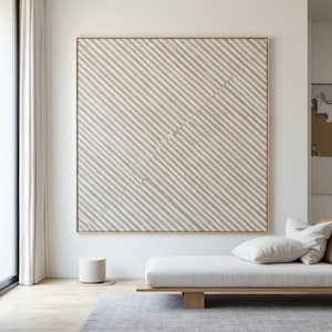 Peut inclure: Grande œuvre d'art carrée abstraite avec un cadre en bois brun clair. L'œuvre présente des rayures diagonales dans des tons de gris et de blanc. L'œuvre est affichée au-dessus d'une méridienne blanche avec des coussins.