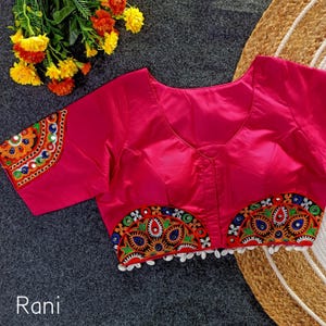 May include: A bright pink blouse featuring a V-neckline and short sleeves. The sleeves and the bottom hem are decorated with semi-circular embroidered patches in orange, green, blue, and white, accented with small white hanging beads.