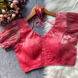 Puede incluir: Una blusa rosa con cuello en V y mangas transparentes con volantes. La blusa tiene un patrón de rayas verticales y un cierre de lazo en la parte posterior del cuello. Cuelga de una percha de madera.
