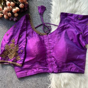 May include: A striking purple blouse with detailed gold embroidery on the sleeves and neckline. The blouse has a V-neck and short sleeves, with a tie closure at the back. The fabric has a subtle sheen, enhancing its elegant look.