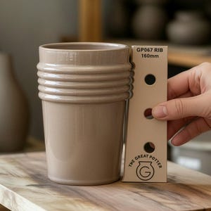 Puede incluir: Un vaso de cerámica beige con detalles acanalados horizontales. Una herramienta de medición muestra que el vaso mide 16 cm de alto. El logotipo de The Great Potter es visible. El vaso está sobre una superficie de madera.