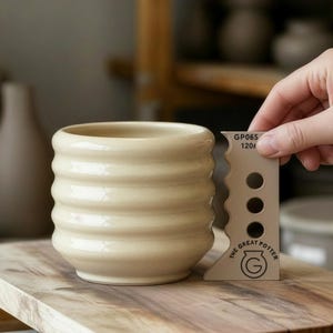 Op de afbeelding: Een beige keramische beker met een geribbeld ontwerp staat op een houten oppervlak. Een gereedschap met de tekst "THE GREAT POTTER" en "GP065 120" wordt naast de beker gehouden. De beker is lichtbeige van kleur.