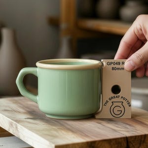 Op de afbeelding: Een groene keramische mok met een beige rand en oor. De mok staat op een houten oppervlak. Een hand houdt een kaart met de tekst "GP049 R 80mm" en "THE GREAT POTTER".