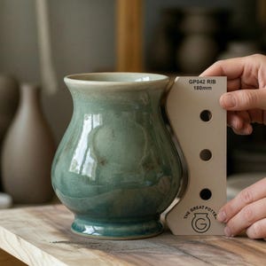 Op de afbeelding: Een handgemaakte, teal-kleurige keramische vaas met een brede body en uitlopende rand. De vaas staat op een houten oppervlak, met een gereedschap met de tekst "GP042 RIB 180mm". De vaas heeft een glanzende afwerking en een craquelé glazuur.