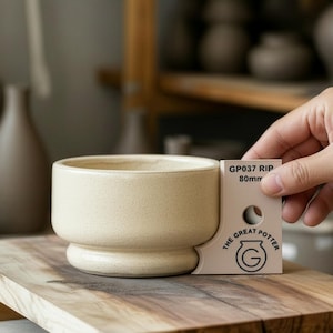 Puede incluir: Un cuenco de cerámica beige con base redondeada y borde ancho. El cuenco está sobre una superficie de madera. Una mano sostiene una herramienta de medición que dice "GP037 RIP 80mm" y "THE GREAT POTTER".