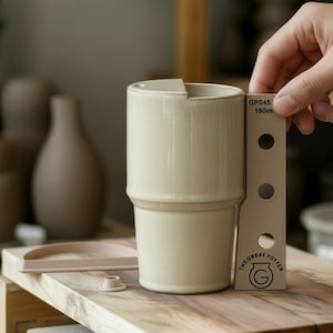 Puede incluir: Una taza de cerámica beige con un diseño escalonado único. La taza se sostiene junto a una regla que mide 16 cm. La taza está sobre una superficie de madera. El texto "THE GREAT POTTER" es visible en la regla.
