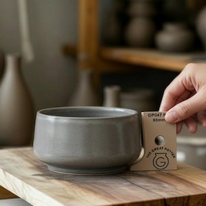 May include: A gray ceramic bowl with a rounded shape sits on a wooden surface. A hand holds a card that reads "GP047 R 80mm" and "The Great Potter" logo. The background shows other pottery items.