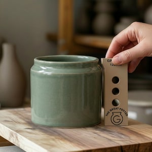 Puede incluir: Un tarro de cerámica verde salvia con una abertura ancha. El tarro es cilíndrico con un borde ligeramente redondeado. Una herramienta de medición beige con el texto "THE GREAT POTTER" se sostiene junto al tarro. El tarro está sobre una superficie de madera.