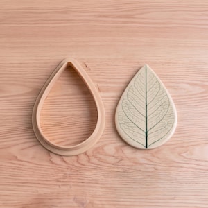 May include: A teardrop-shaped cookie cutter with a matching stamp featuring a detailed leaf vein pattern. The cutter is an open frame, while the stamp is a solid piece with a dark green central vein and fine lines radiating outward.