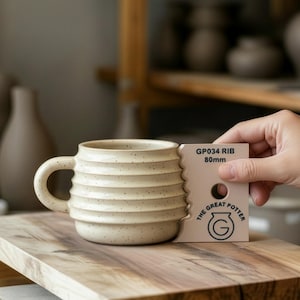 Puede incluir: Una taza de cerámica beige con estrías horizontales y un asa curva sobre una superficie de madera. Una mano sostiene una herramienta rosa con la inscripción "GP034 RIB 80mm" y "THE GREAT POTTER".