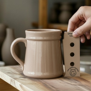 May include: A beige ceramic mug with a handle, on a wooden surface. A hand holds a beige card with the text "GP06 140" and "THE GREAT POTTER" logo. The mug has a ribbed design and a wide rim.