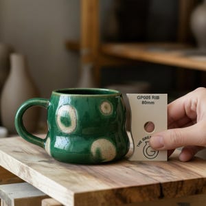 May include: A white ceramic mug with a black handle and the text "But First, Coffee" in a bold, black font. The mug is set against a blurred background, suggesting a cosy setting.