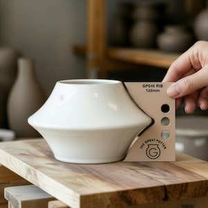May include: A white ceramic vase with a unique, flared shape. The vase is displayed on a wooden surface. A hand holds a color swatch card that reads "GP055 RIB 120mm" with color samples and the logo "The Great Potter".