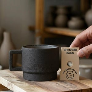 以下が含まれることがあります： 黒いセラミックマグカップ。表面はざらざらしており、四角いハンドルが付いています。マグカップは木製の表面に置かれています。「GP012 RIB 80mm」と書かれたベージュ色の長方形のツールがマグカップの隣にあります。