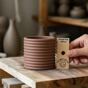 Può includere: Un vaso in ceramica marrone con scanalature orizzontali. Il vaso cilindrico è mostrato con una scheda beige che indica "GP009 RIB 100mm". La scheda mostra anche un logo e "THE GREAT POTTER". Il vaso misura circa 10 cm di altezza.