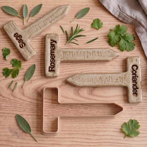 May include: Three beige ceramic garden markers with the words Sage, Rosemary, and Coriander embossed in dark brown, accompanied by one matching beige plastic T-shaped cookie cutter, surrounded by scattered fresh green herb leaves.
