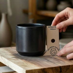 Puede incluir: Una taza de cerámica negra mate con forma cilíndrica y base ligeramente ensanchada. La taza está sobre una superficie de madera. Una tarjeta beige junto a la taza indica "GP068 RI 100mm". La taza mide unos 10 cm de alto.