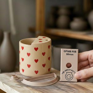 Könnte beinhalten: Ein beigefarbener Keramikbehälter mit roten Herzmustern, einem passenden Deckel und einem kleinen, runden Teil. Ein rechteckiges Werkzeug mit dem Text "GP006 RIB 80mm" wird in der Nähe gehalten. Der Behälter hat eine gewölbte Oberseite.