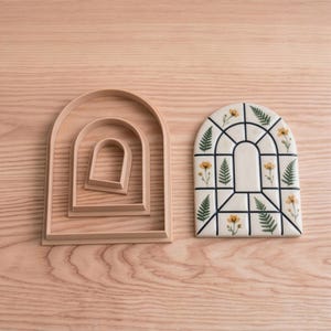 Könnte beinhalten: Ein Set aus drei ineinander verschachtelten, bogenförmigen Ausstechformen in hellem Beige, platziert neben einer bogenförmigen Keramikfliese mit einem Gittermuster, das mit kleinen gelben Blumen und grünen Farnblättern auf cremefarbenem Grund verziert ist.