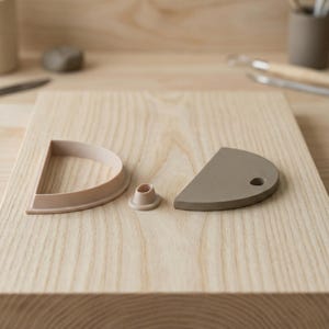 May include: A collection of pottery tools, including a half-circle shaped clay cutter with a hole, a small cylindrical tool, and a D-shaped cutter. The tools are in shades of beige and gray, arranged on a light wood surface.