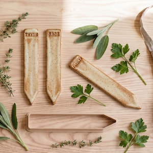 May include: Three rectangular ceramic plant markers with pointed ends, featuring the stamped text THYME, SAGE, and PARSLEY. The markers have a speckled cream glaze. A single empty plastic marker mould of the same shape is positioned below.