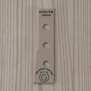 May include: A beige pottery rib tool with three circular holes and the text "GP045 RIB 160mm". The tool also features a logo that says "THE GREAT POTTER". The tool is set against a light wood grain background.