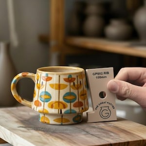 May include: A patterned ceramic mug with a cream base and a retro floral design in orange, yellow, and blue. The mug has a handle and a wide base. A beige tool with the text "GP002 RIB 100mm" is next to the mug.