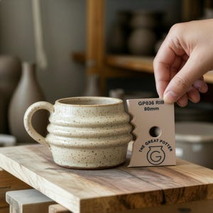 Puede incluir: Una taza de cerámica beige moteada con un diseño acanalado y un asa curva se encuentra sobre una superficie de madera. Una herramienta con el texto "GP036 RIB 80mm" se sostiene cerca. La taza es parte de una colección de cerámica.