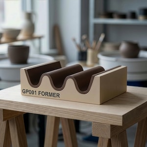 May include: A beige and brown pottery former with a wavy design. The former has three curved slots and the text "GP001 FORMER" printed on the front. It sits on a wooden table in a pottery studio.