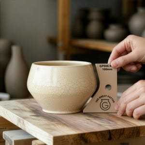 May include: A cream-coloured ceramic bowl with a crackle glaze finish sits on a wooden surface. A tan-coloured tool with the text "THE GREAT POTTER" and "GP056 R 100mm" is held next to the bowl. The bowl has a wide opening and a rounded shape.