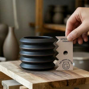 May include: A black ceramic planter with horizontal ridges and bronze accents. The planter is on a wooden surface. A hand holds a tool that reads "GP024 100mm" and "The Great Potter".