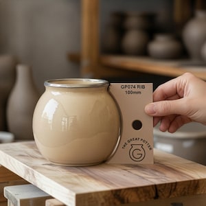Może przedstawiać: Beżowy ceramiczny garnek o zaokrąglonym korpusie i metalowej krawędzi. Beżowe narzędzie garncarskie jest umieszczone przy boku garnka, z napisem GP074 RIB 100mm oraz logo The Great Potter z okrągłą ikoną G i małym otworem.