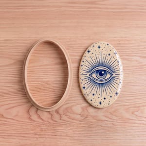 May include: An oval cookie cutter with a light beige frame and a matching oval plaque featuring a dark blue graphic of a central eye surrounded by radiating lines and small star shapes on a cream-coloured background.