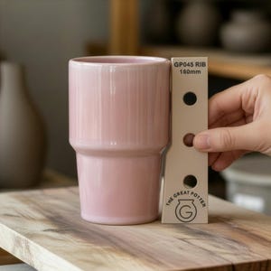 Può includere: Una tazza in ceramica rosa chiaro con forma cilindrica e base a gradini. Uno strumento di misurazione indica "GP045 RIB 160mm". Il logo The Great Potter è visibile. La tazza è su una superficie di legno.