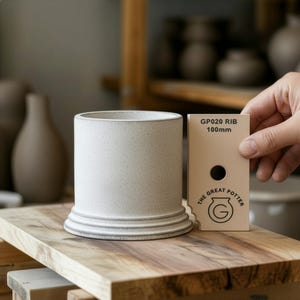 May include: A cylindrical ceramic pot in speckled off-white, with a tiered base. The product is displayed on a wooden surface. A card held nearby reads "GP020 RIB 100mm" and "THE GREAT POTTER".