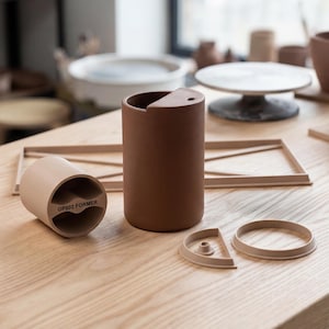 以下が含まれることがあります： 上部が湾曲し小さな穴のある茶色のセラミックタンブラー。テキスト「GP002 FORMER」の付いたベージュのセラミックピースの隣にあります。追加のベージュのセラミックピースと木製のフレームも木製の表面にあります。