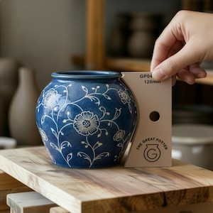 Könnte beinhalten: Eine runde, blaue Keramikvase mit weißen Blumenmustern. Die Vase hat einen dunkelblauen Rand und einen Durchmesser von etwa 12 cm. Die Vase steht auf einer Holzoberfläche. Eine Hand hält ein Messwerkzeug neben der Vase.