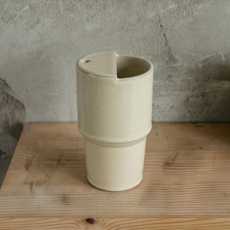 May include: A cream-colored ceramic tumbler with a unique design. The top half of the tumbler has a built-in lid with a small hole. The bottom half has a slightly wider base. The tumbler sits on a wooden surface.